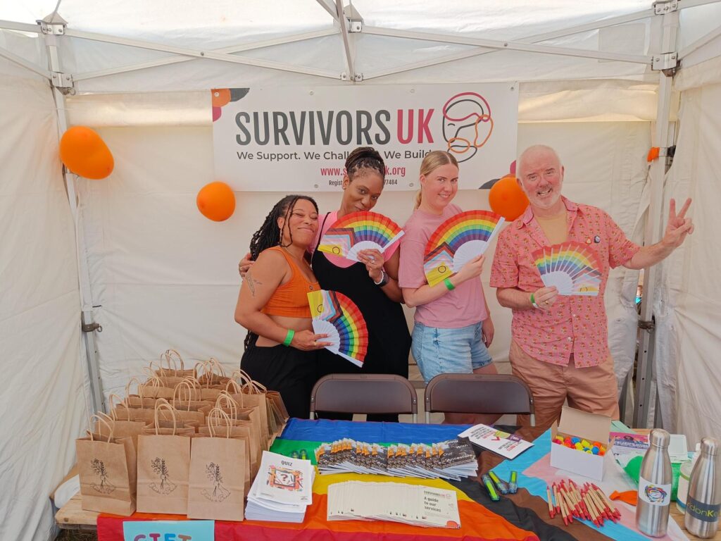 Four SurvivorsUK colleagues with Pride fans at the SurvivorsUK stall. On the table there are goody bags, poetry books, leaflets and colourful pens