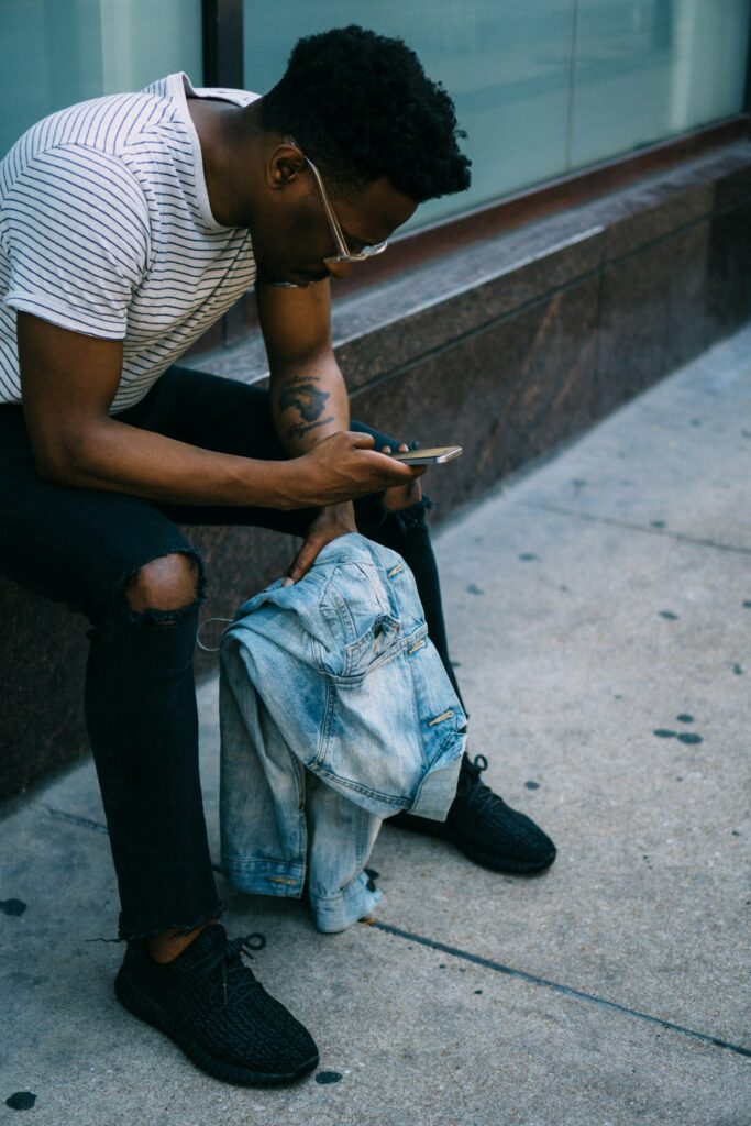 A young black man sits down at a bench with his phone and talks to the webchat helpline. He has short black hair and wears glasses, a striped t-shirt and jeans.