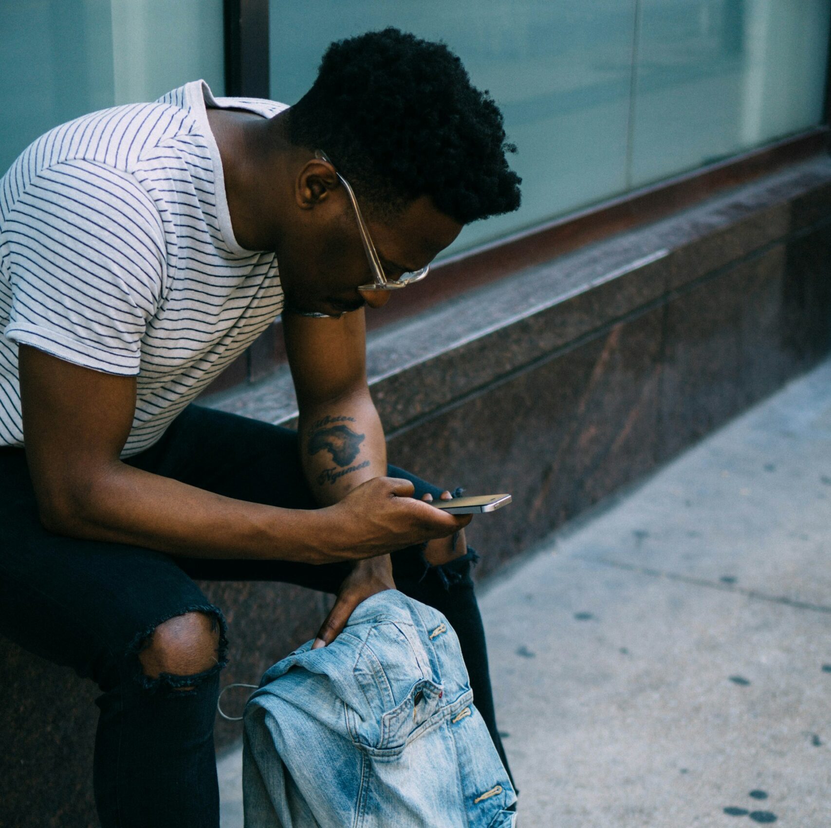 A young black man sits down at a bench with his phone and talks to the webchat helpline. He has short black hair and wears glasses, a striped t-shirt and jeans.